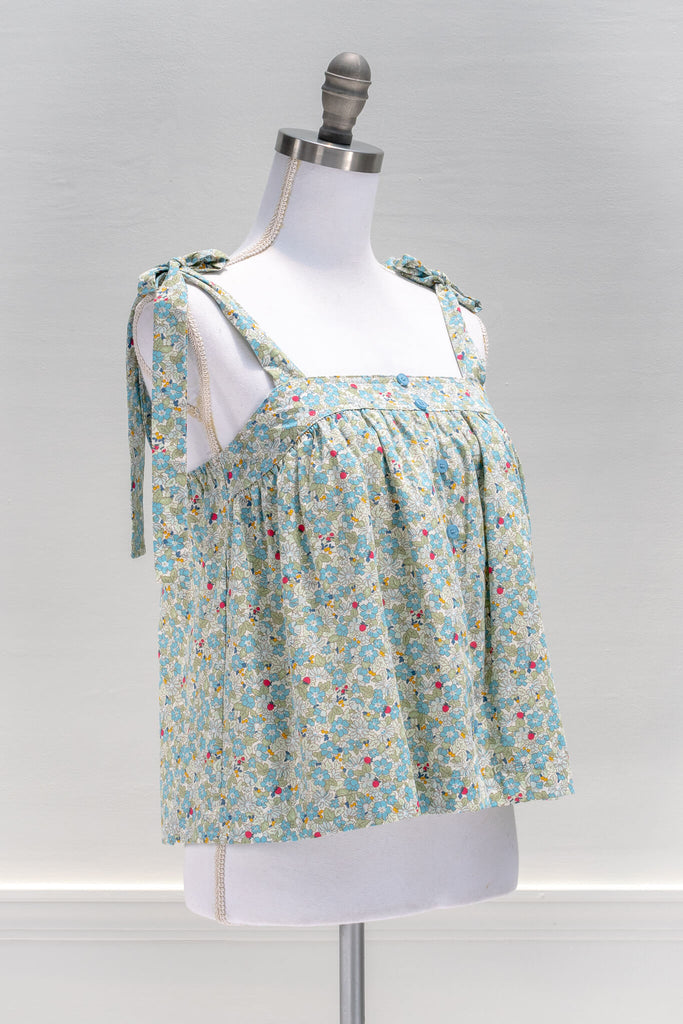 The feminine aesthetic Watercolorist camisole features a blue and yellow floral print, tie-style shoulders, a button-up front, and a cute babydoll smock-style cut. amantine clothes - quarter view 