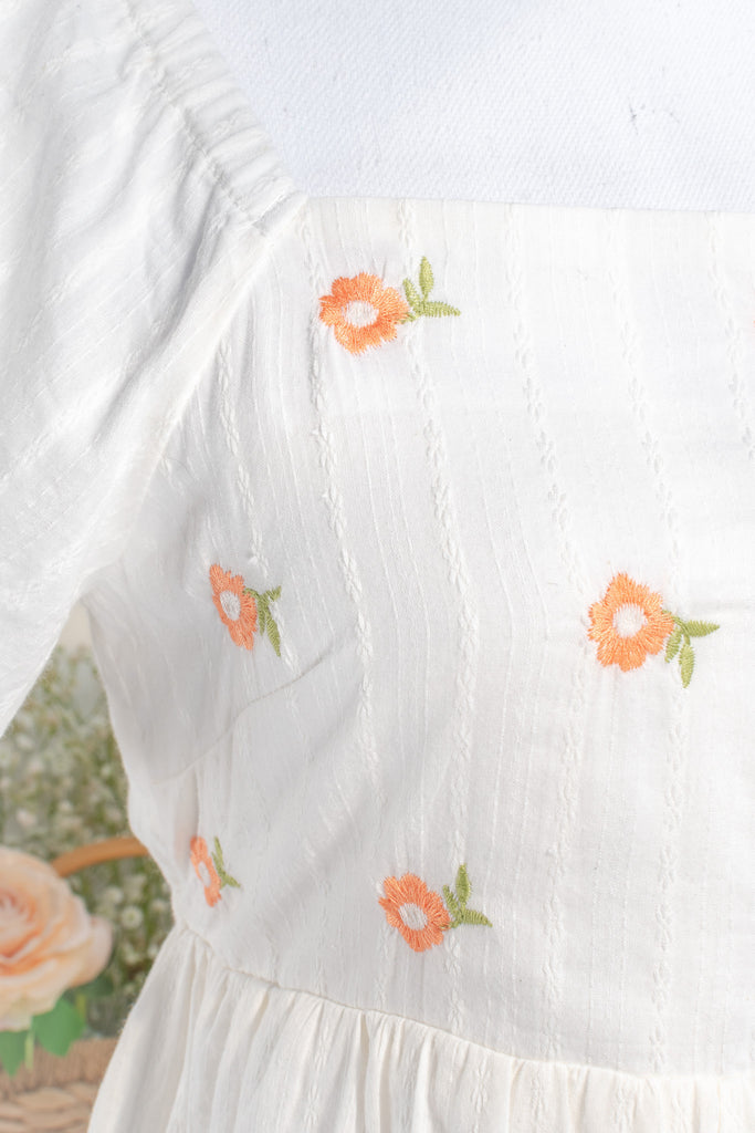 close-up view of white cottagecore dress fabric highlighting intricate orange floral embroidery with penny-sized flowers adding a delicate and vintage-inspired detail.