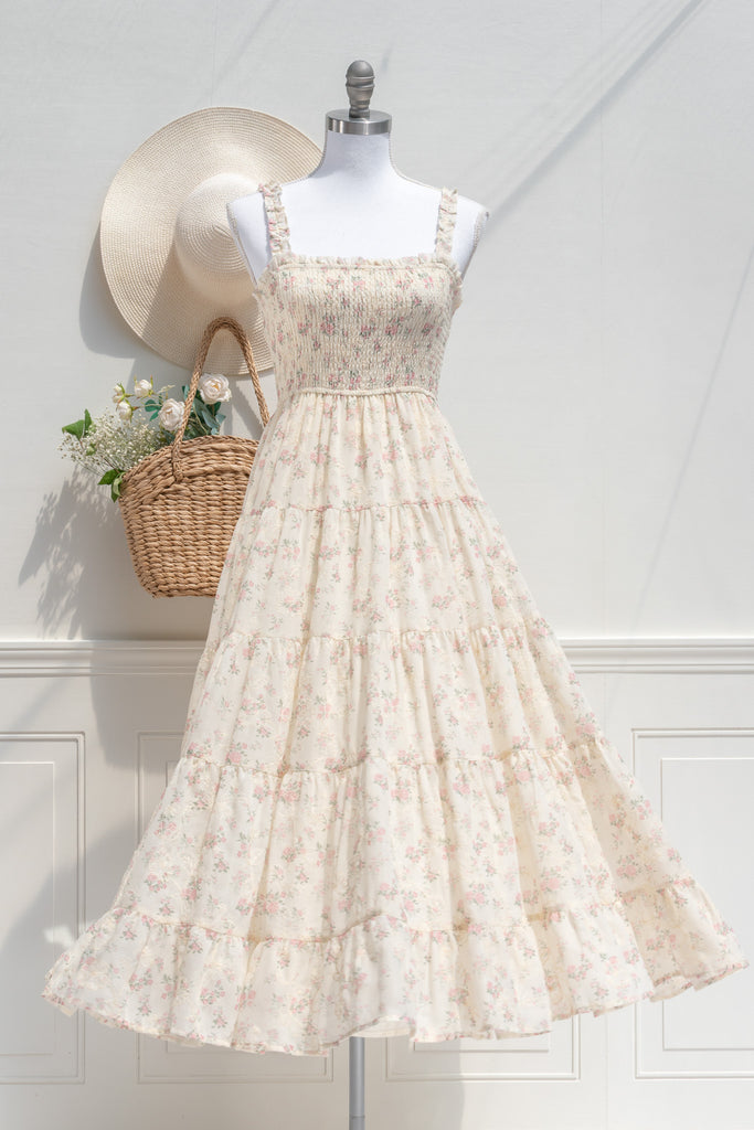 Front view of a cream-colored cottagecore sundress with pink floral print, smocked bodice, ruffled straps, and a flowing tiered skirt, styled with a straw hat and wicker basket.
