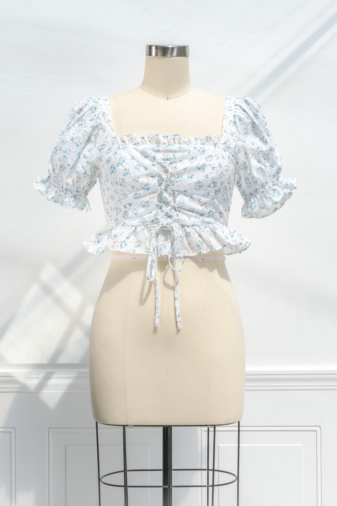 feminine clothing - photo shows a feminine blouse, with a square neckline, puff sleeves, ruffled waist, and cropped lenght with a light blue on white floral print - front view. 