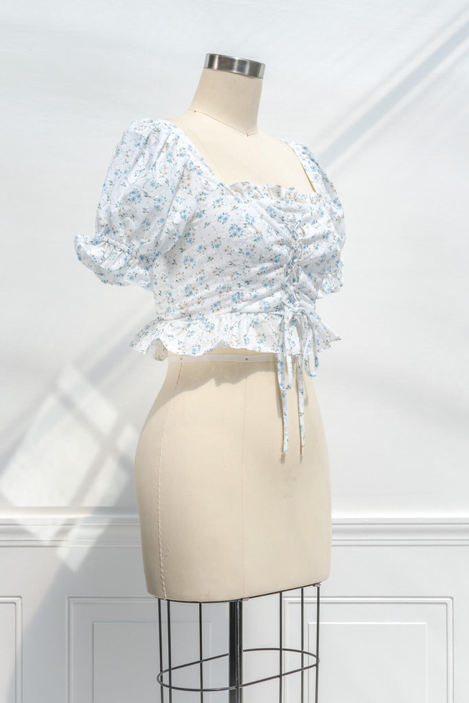 feminine clothing - photo shows a feminine blouse, with a square neckline, puff sleeves, ruffled waist, and cropped lenght with a light blue on white floral print - quarter side view. 