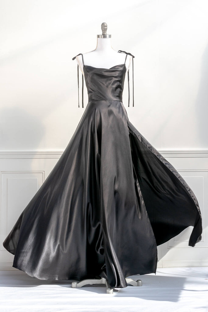 french elegant long black gown - made of a silky and shiny black fabric, with spaghetti straps, and a french feminine cowl neckline. features a side slit. shot billowing in the wind view - amantine.   