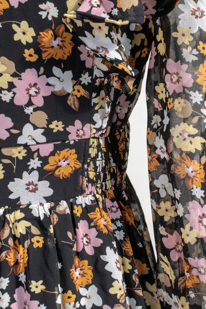 cottagecore and cottage core dresses in a vintage and french style cut. a v neck, long sleeve, midi floral dress. it features a black chiffon with yellow, white, and orange little flowers throughout. up close view showing seams, and ruching around waist. amantine cottagecore dresses.