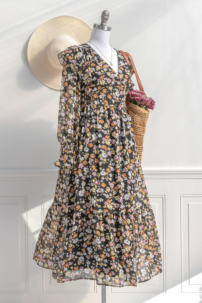 cottagecore and cottage core dresses in a vintage and french style cut. a v neck, long sleeve, midi floral dress. it features a black chiffon with yellow, white, and orange little flowers throughout. styled with a french bag and french hat view. amantine cottagecore dresses.