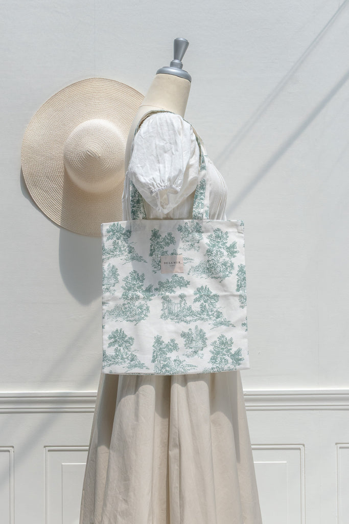 Mannequin wearing a white dress with a green floral patterned bag, next to a straw hat on a white wall.