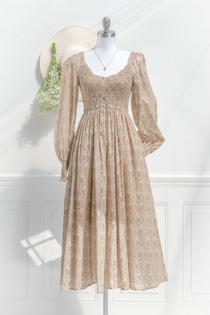 prairie dress in a fall fashion style. round neckline, long sleeves, smocked bodice, long skirt. Taupe brown cotton fabric with floral details in in cream and brown. front view with sleeve in pocket. amantine 