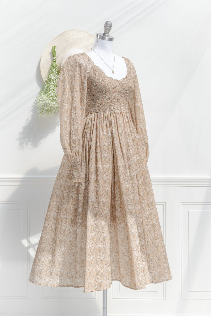 prairie dress in a fall fashion style. round neckline, long sleeves, smocked bodice, long skirt. Taupe brown cotton fabric with floral details in in cream and brown. 3/4 side view. amantine 