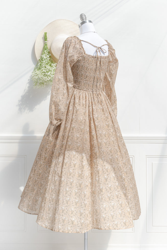 prairie dress in a fall fashion style. round neckline, long sleeves, smocked bodice, long skirt. Taupe brown cotton fabric with floral details in in cream and brown. back 3/4 view. amantine 